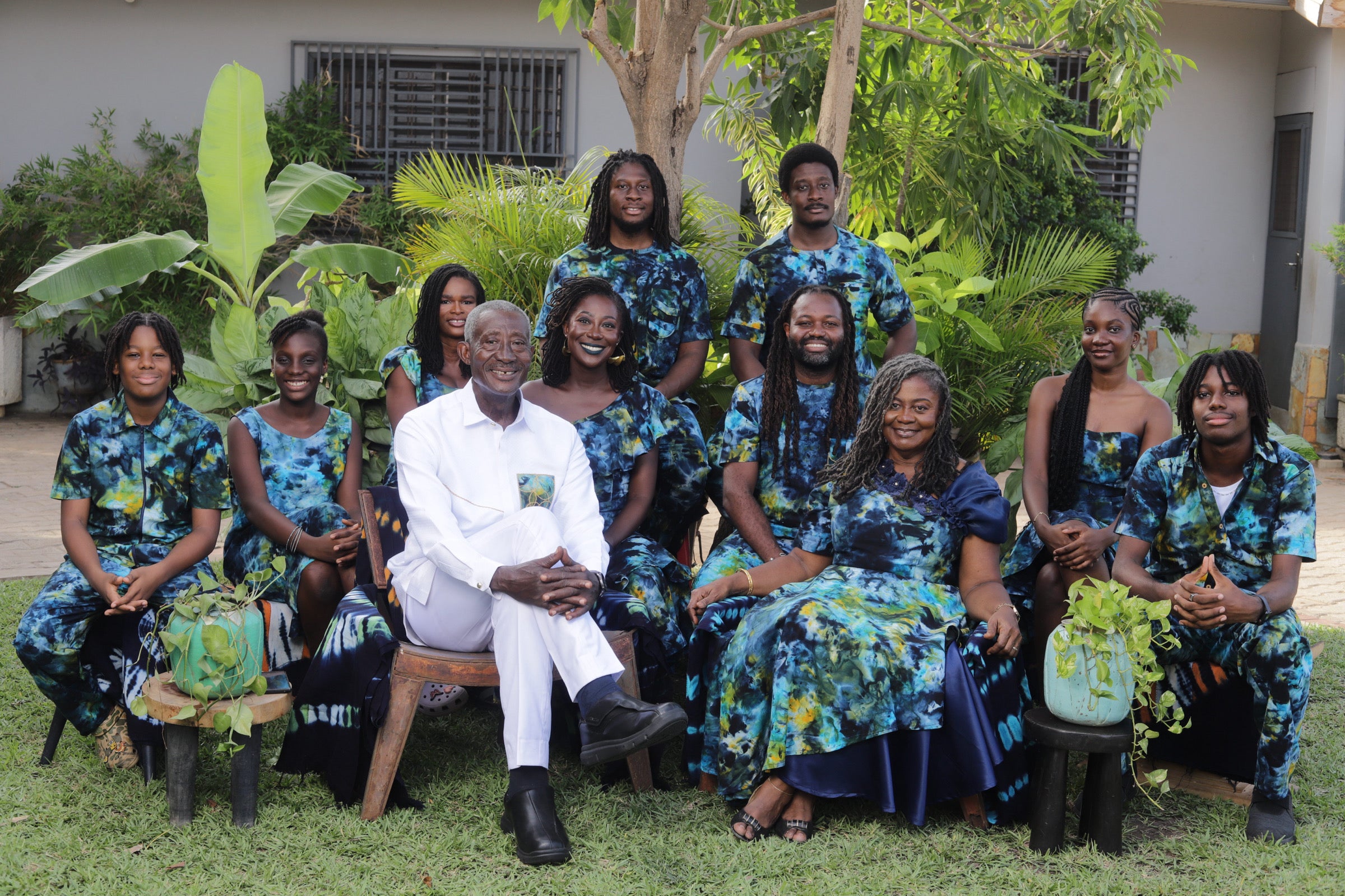 Family group portrait in matching blue green Tampoori Bubblegum tie-dye floral outfits with a central figure, the birthday celebrant in a white outfit 70th birthday outfit seated outdoors and surrounded by greenery
