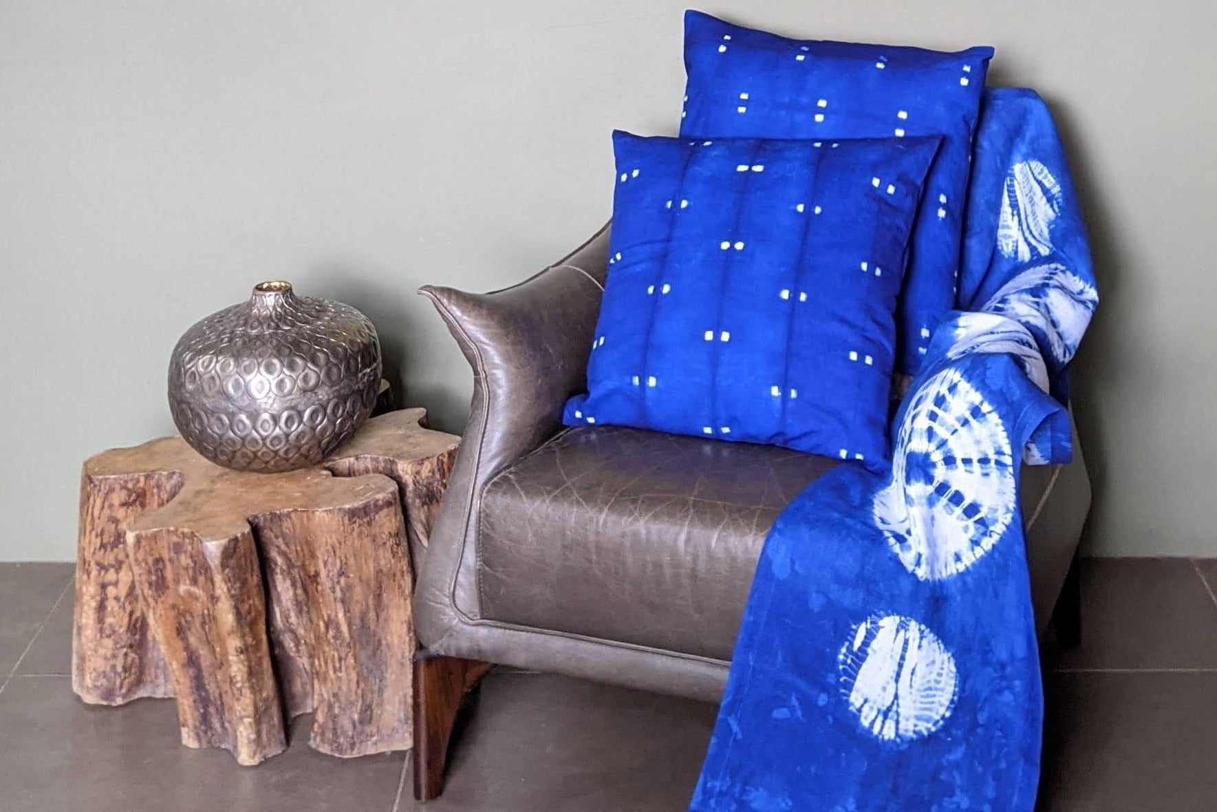 Blue patterned hand-dyed batik tie dye blanket and pillows on a brown chair against a gray wall.