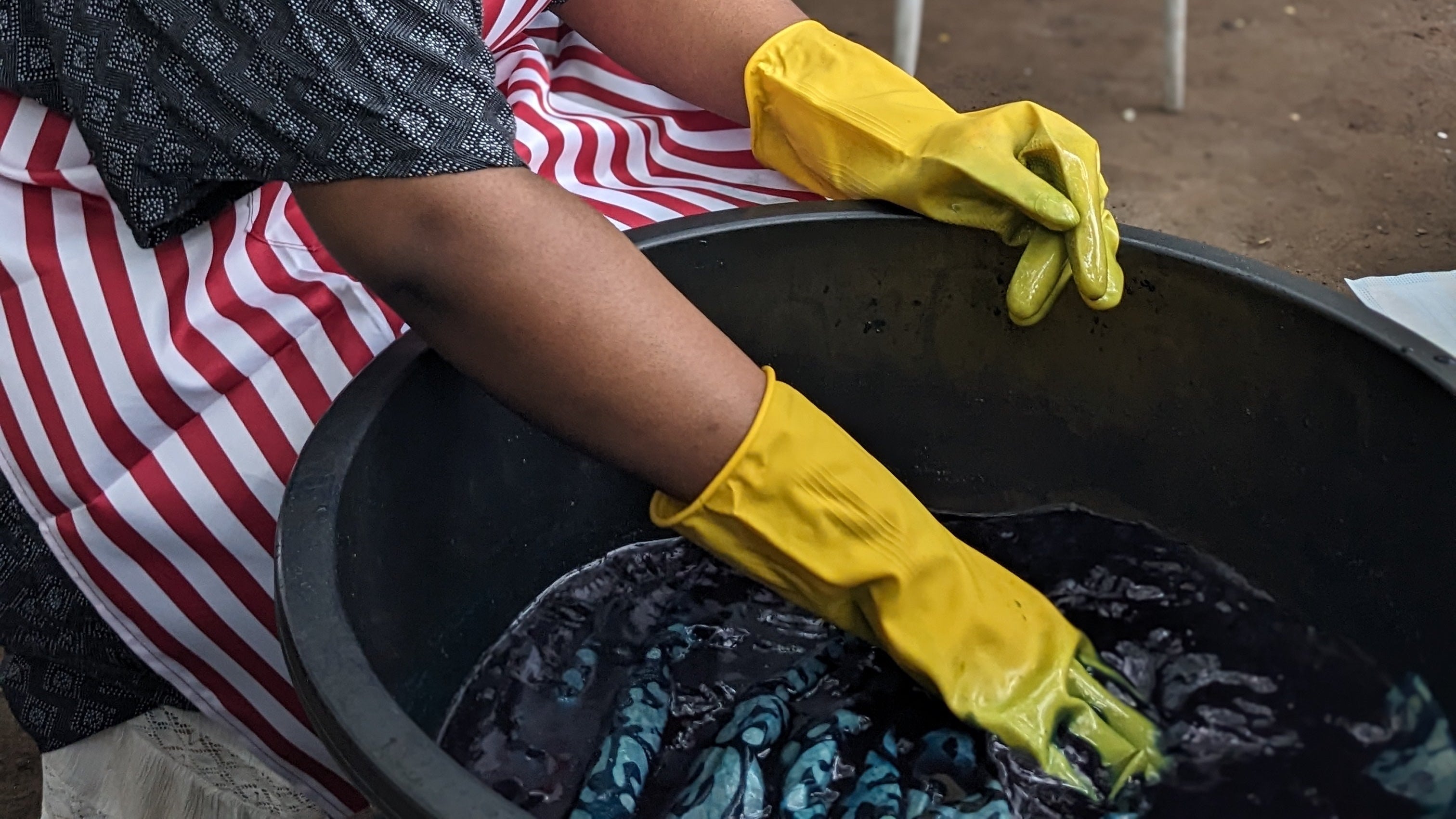 Tampoori textile Artisan pouring dye over wax-patterned fabric in a large black vat.