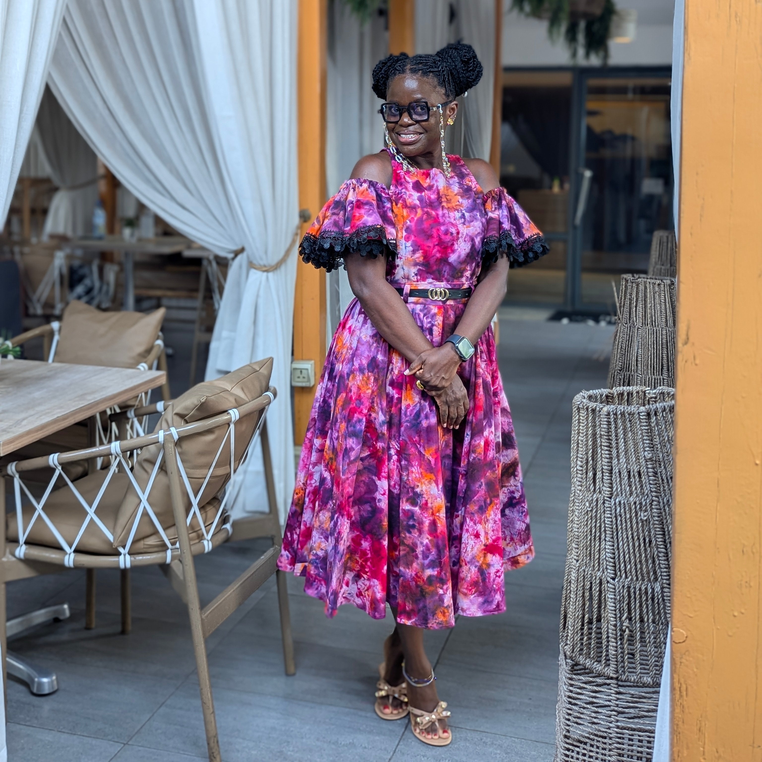 Woman in a colorful Tampoori Bubblegum cherry tie dye dress standing in an outdoor setting with tables and chairs.