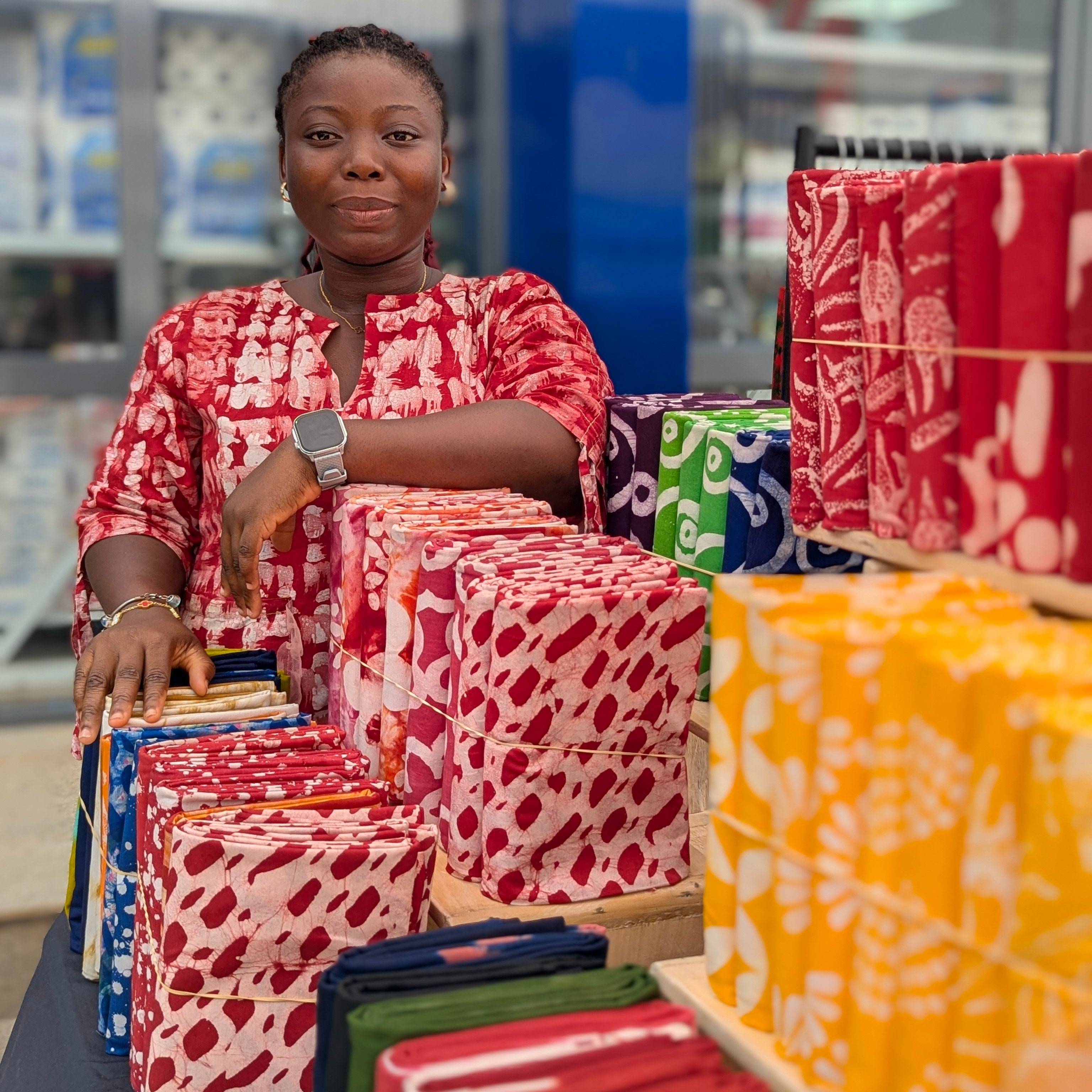Woman in a red patterned outfit standing behind a display of colorful fabric tampoori-batik-shop-labone-accra.
