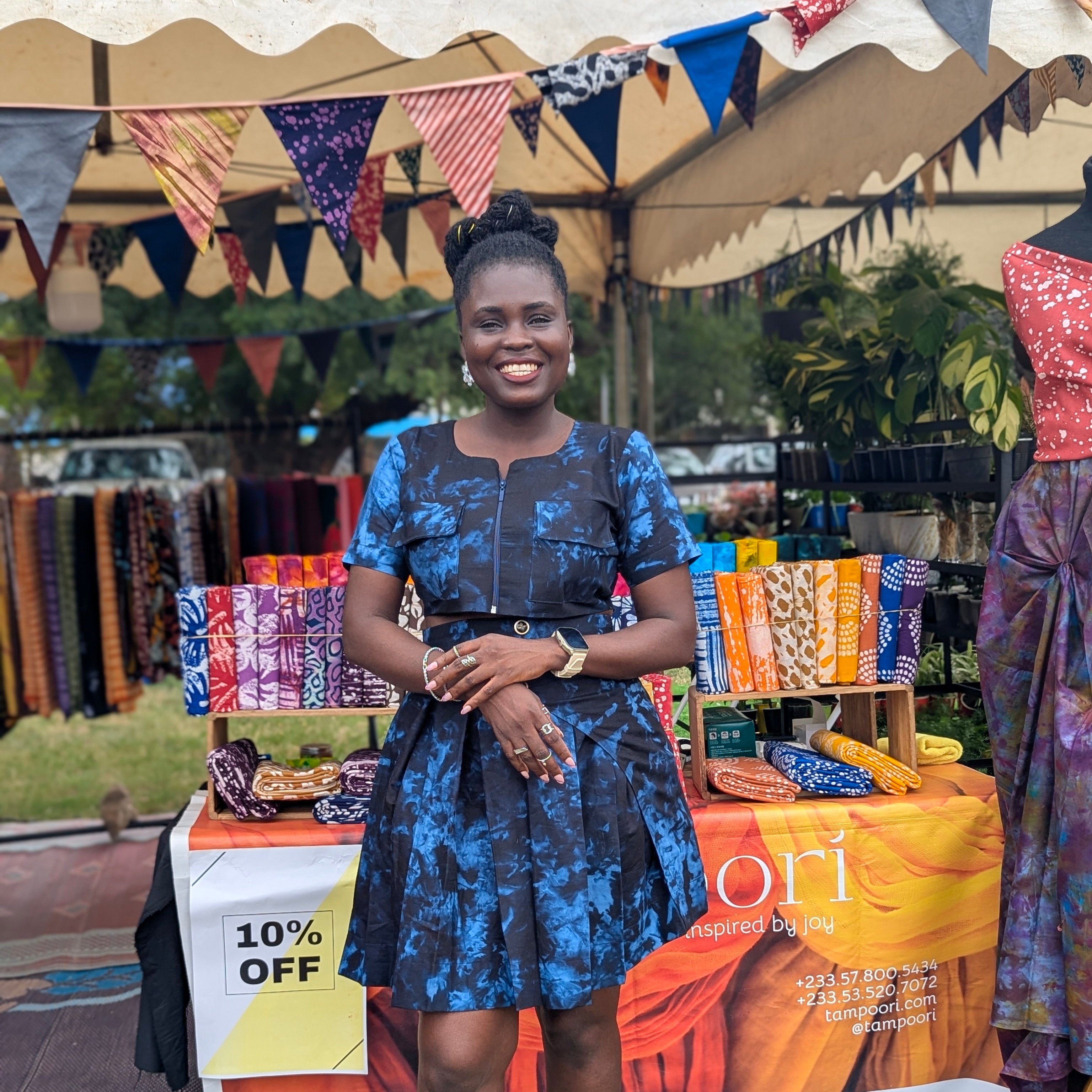 Woman wearing batik tie-dye made by hand by Tampoori in Bubblegum pattern standing in front of a colorful stall with Tampoori branded pop up at a fair